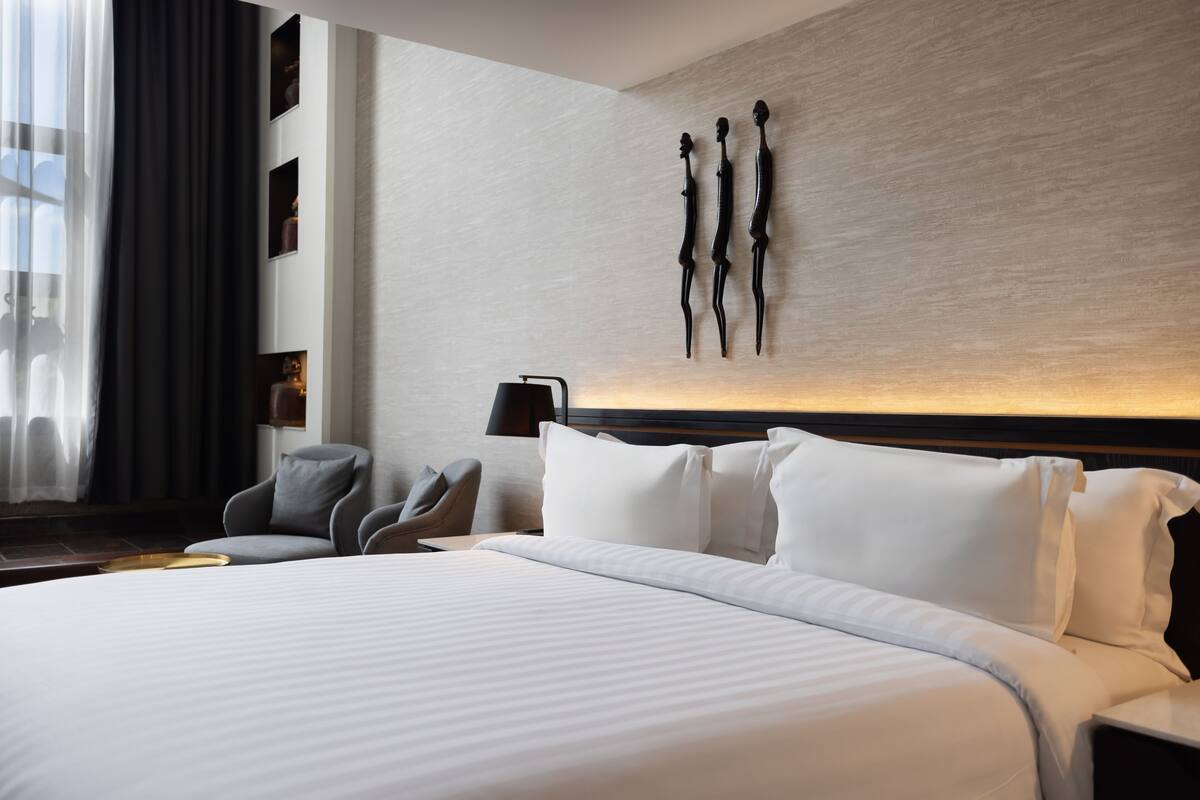 Moderni hotellihuone, jossa on suuri sänky, oleskelualue tuoleineen, seinälle ripustettu taideteos sekä yöpöytä lampilla.