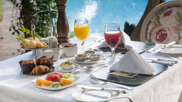 Disposição de café da manhã com croissants, frutas e bebidas em uma mesa perto de uma piscina.