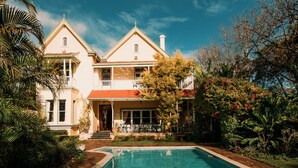 Um edifício de dois andares com piscina, cercado por vegetação exuberante e um céu azul límpido.