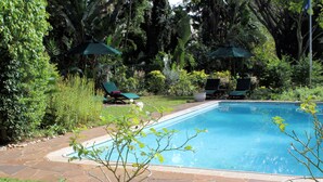 Piscina com borda de pedra, rodeada por grama e plantas. Há duas espreguiçadeiras e dois guarda-sóis.