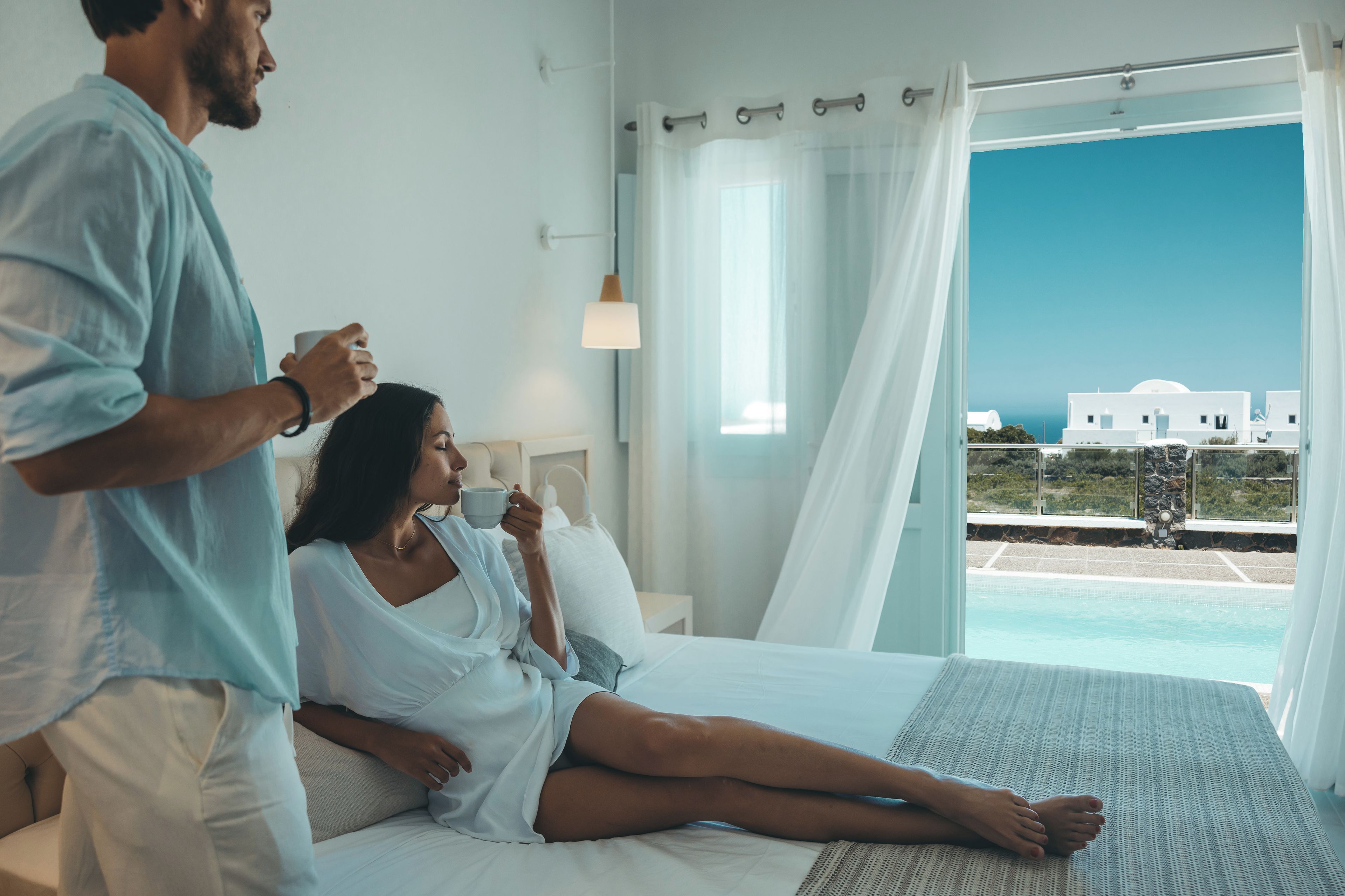 A bedroom with a bed, a person sitting on the bed, and another person standing beside it.