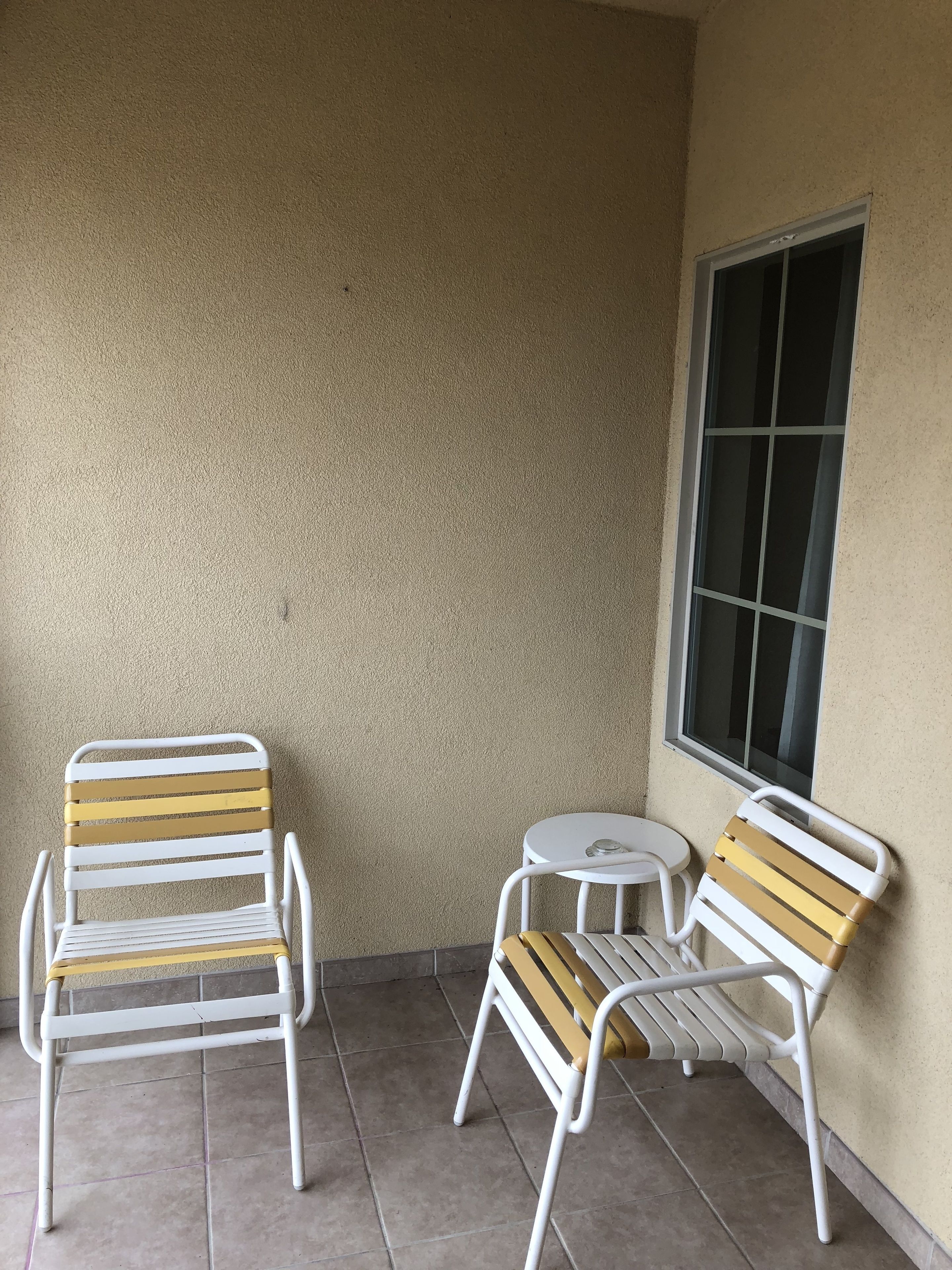 A patio with two white metal chairs and a small round table, against a textured beige wall with a window.