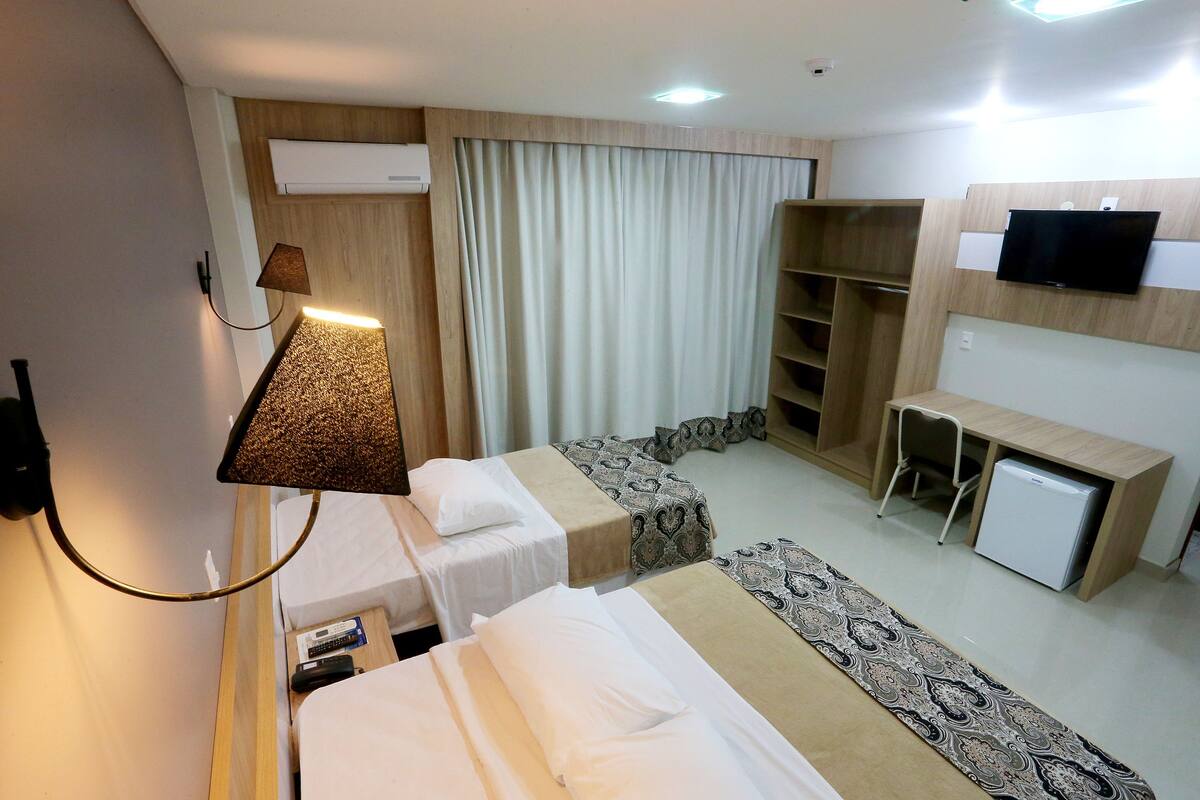 Quarto de hotel com duas camas, uma escrivaninha, televisão, mini-geladeira e guarda-roupa.
