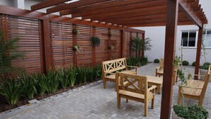 Un patio con mobiliario de madera, una pérgola y una valla decorativa con plantas colgantes.
