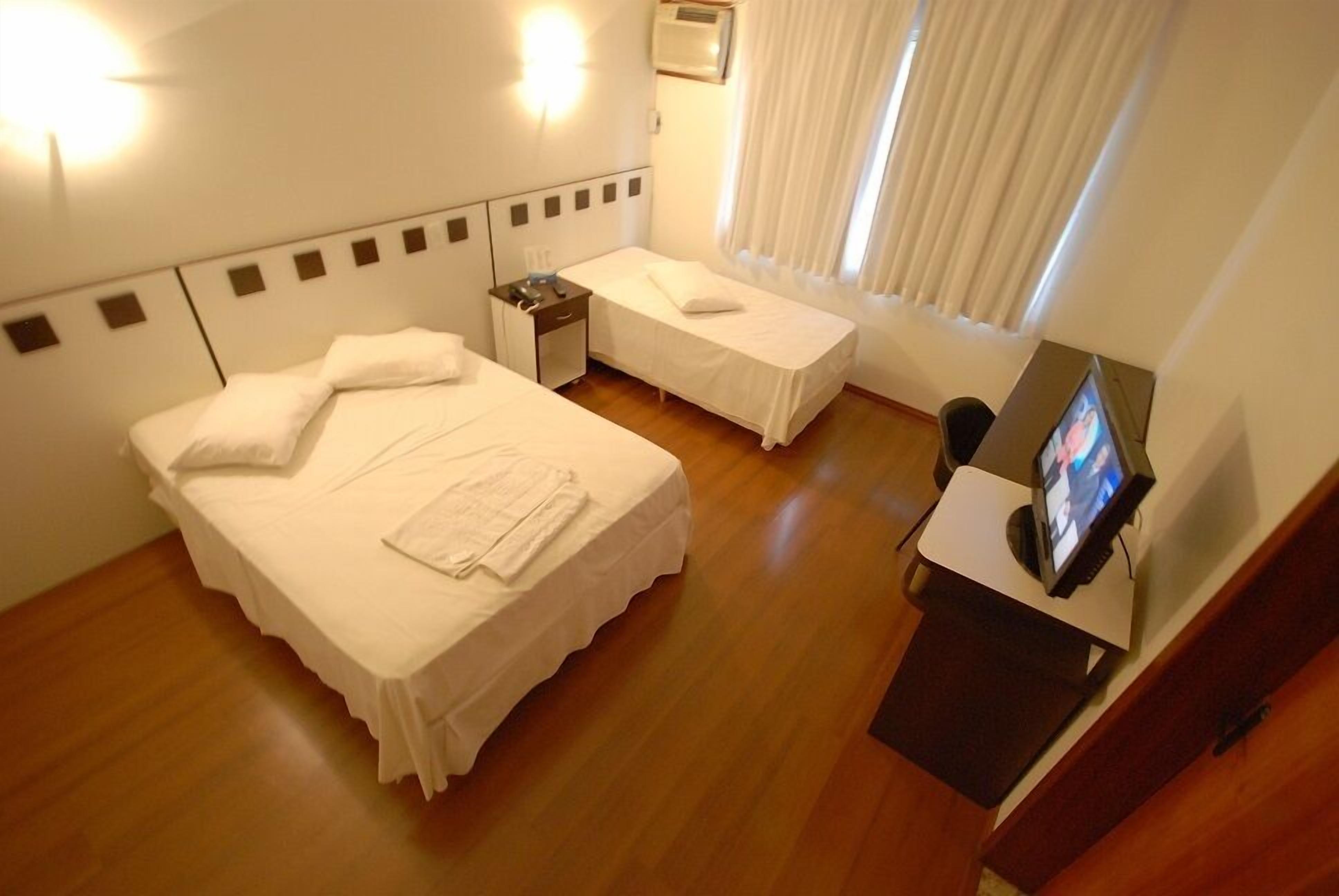 Una habitación de hotel con dos camas, un escritorio con televisor y una ventana con cortinas.