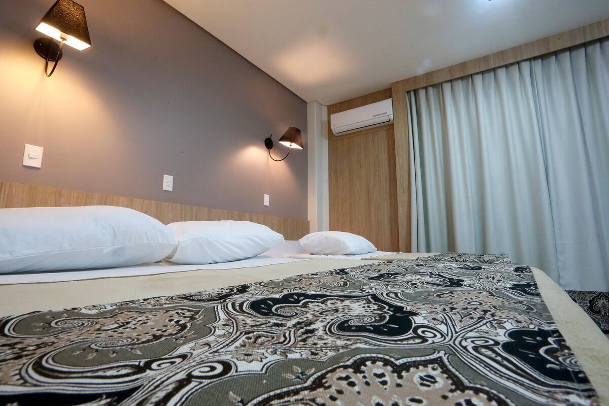 Quarto de hotel com cama, duas luminárias na parede e janela com cortinas.