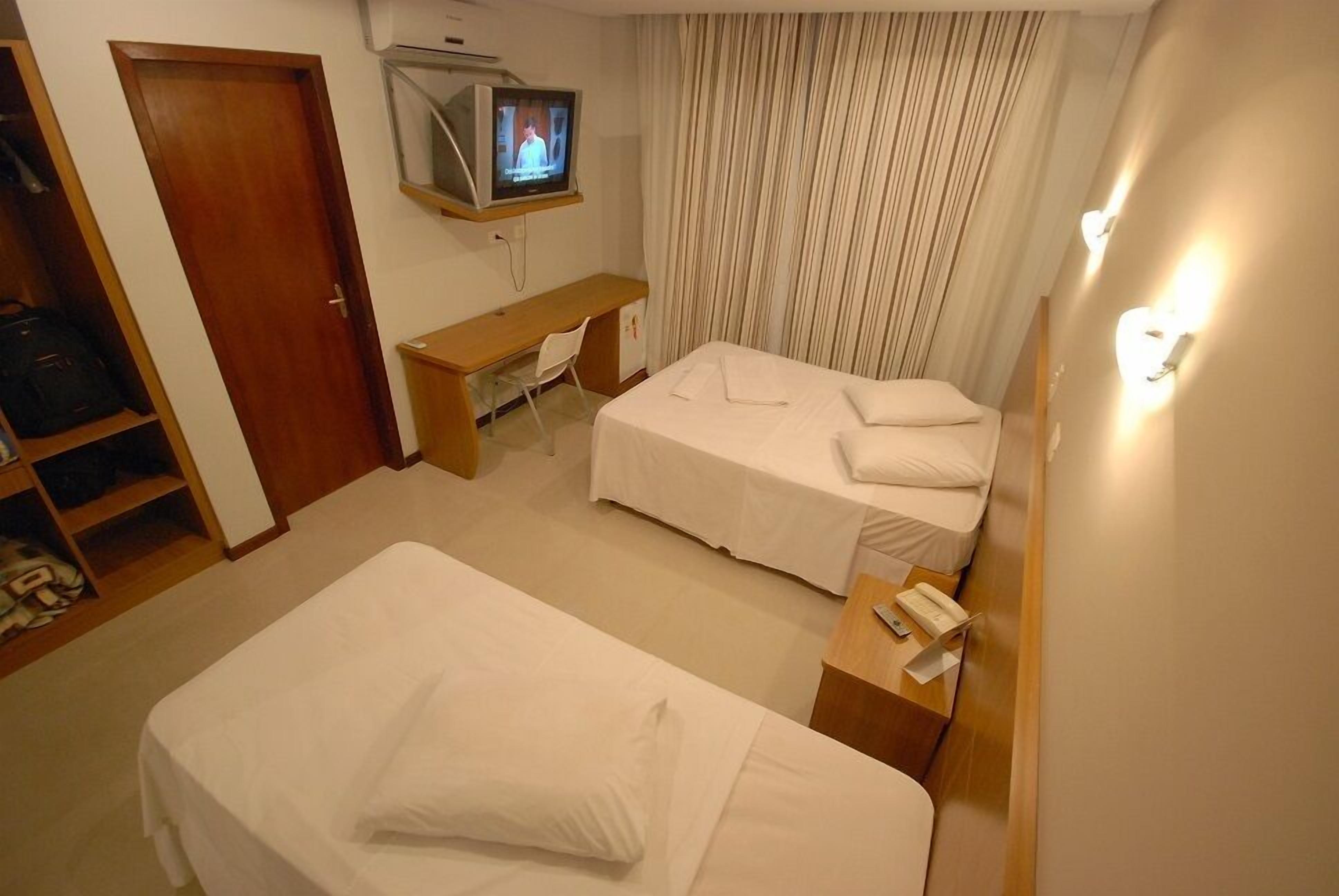 Quarto de hotel com uma cama, uma escrivaninha, televisão e um guarda-roupa.