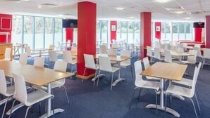 A modern dining area with tables and chairs, large windows, and red accents. - Travelodge Heathrow Terminal 5 (Slough)
