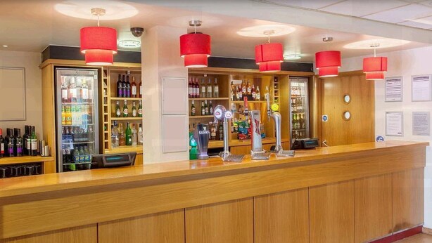 A bar area with wooden counters, red pendant lights, and shelves stocked with various bottles. - Travelodge Heathrow Terminal 5 (Slough)