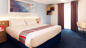 A modern hotel room with a large bed, a nightstand, a bedside table, a chair, and a window with curtains. - Travelodge Heathrow Terminal 5 (Slough)