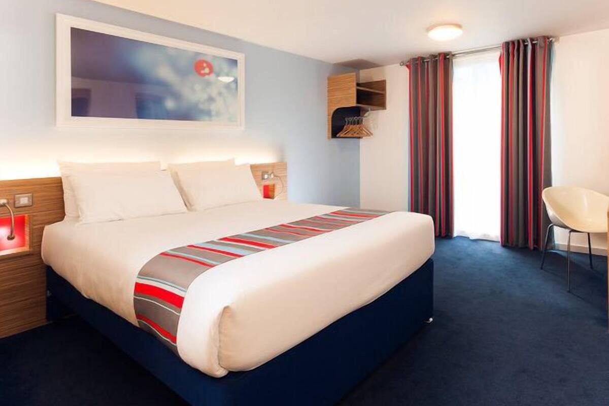 A modern hotel room with a large bed, a nightstand, a bedside table, a chair, and a window with curtains.