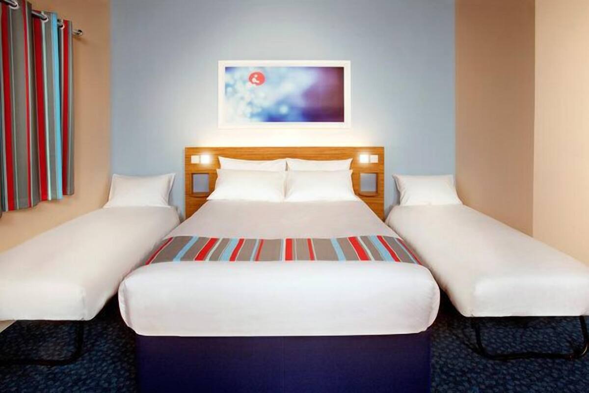 A hotel room with a large bed, two single beds, a headboard with lights, a framed picture on the wall, and a striped blanket.