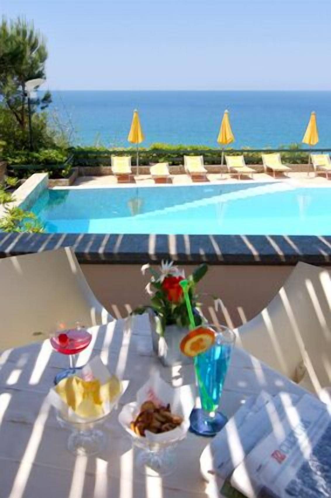A poolside table with drinks and snacks, overlooking a pool with lounge chairs and umbrellas.