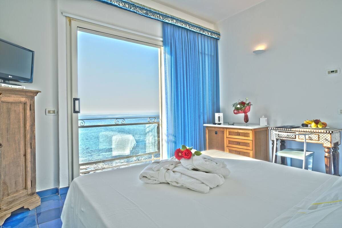 A hotel room with a bed, a wooden cabinet, a desk with a chair, a small table with a vase of flowers, and a view of the sea through a large window.