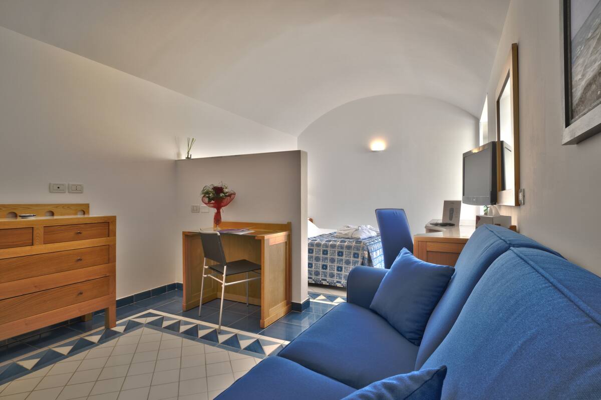 A compact room with a blue sofa, wooden desk, and a bed with patterned bedding.