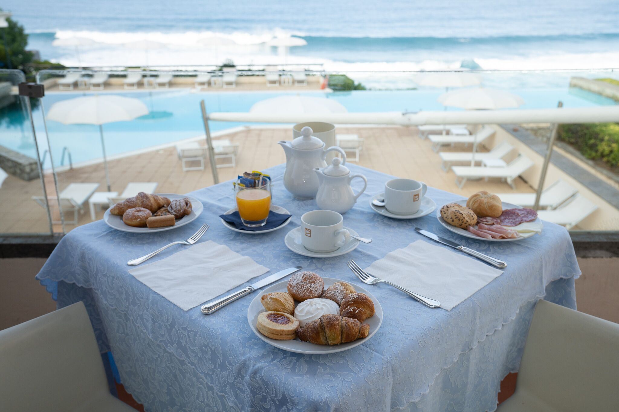 Mesa de desayuno con croissants, pasteles y una taza de café, con vistas a una piscina y una playa.