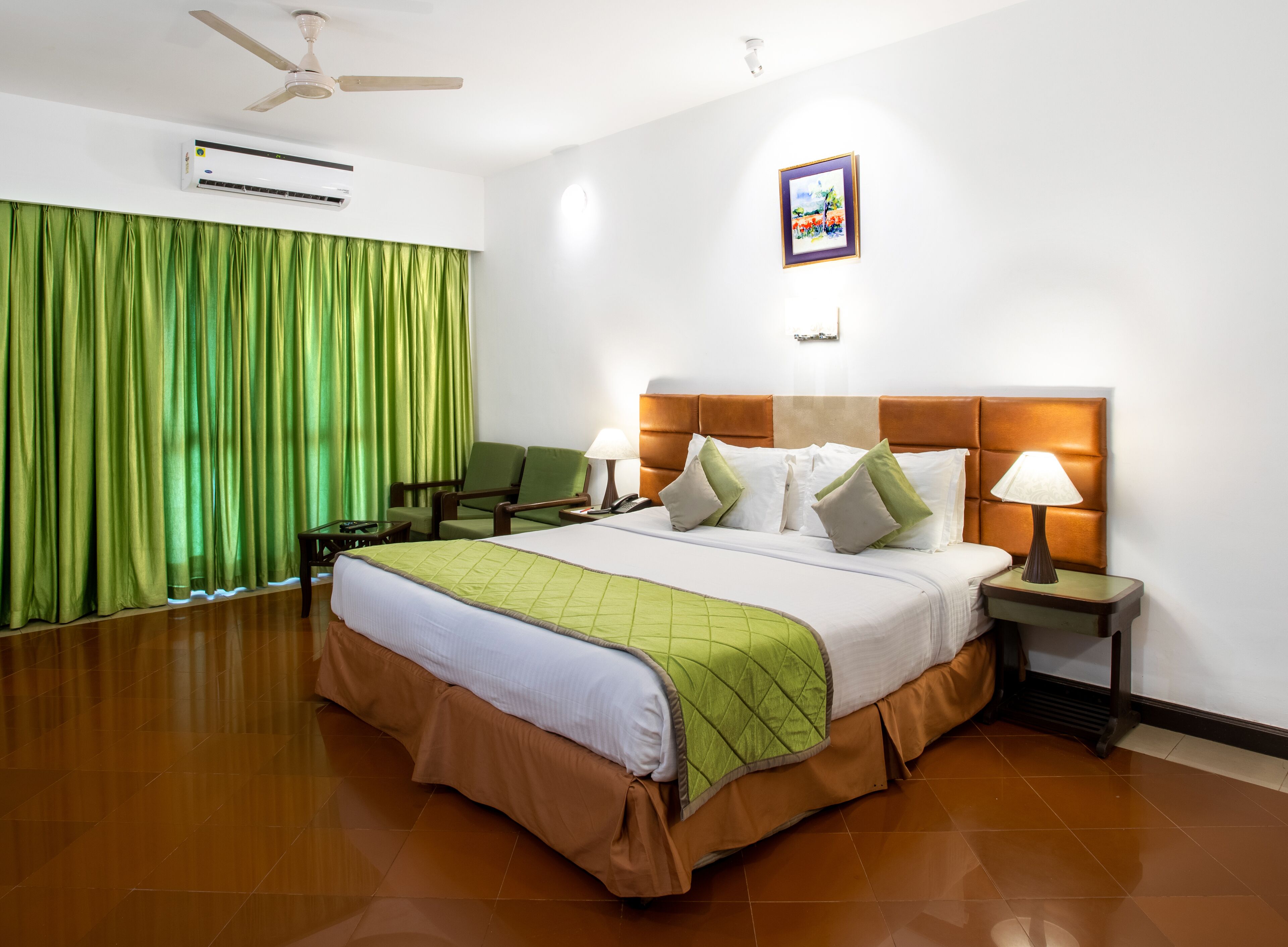 A hotel room with a large bed, a green curtain, a ceiling fan, an air conditioner, a nightstand with a lamp, and a framed picture on the wall.