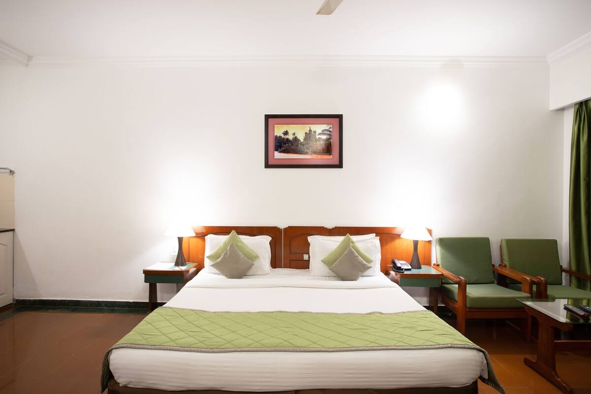 Quarto de hotel com uma cama, duas mesas de cabeceira com luminárias, duas cadeiras, uma cortina verde e um quadro na parede.