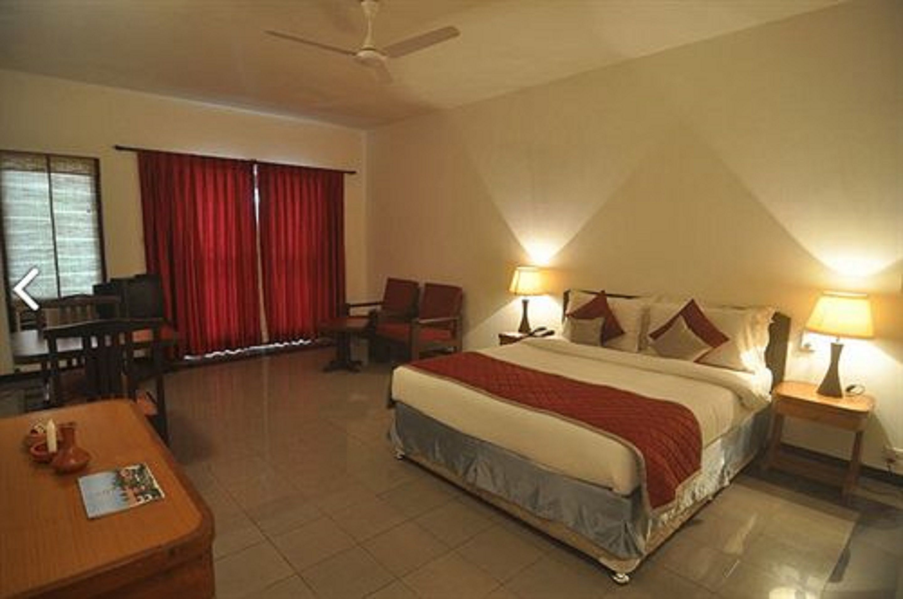 A hotel room with a bed, two bedside tables, a desk, a chair, and a window with red curtains.