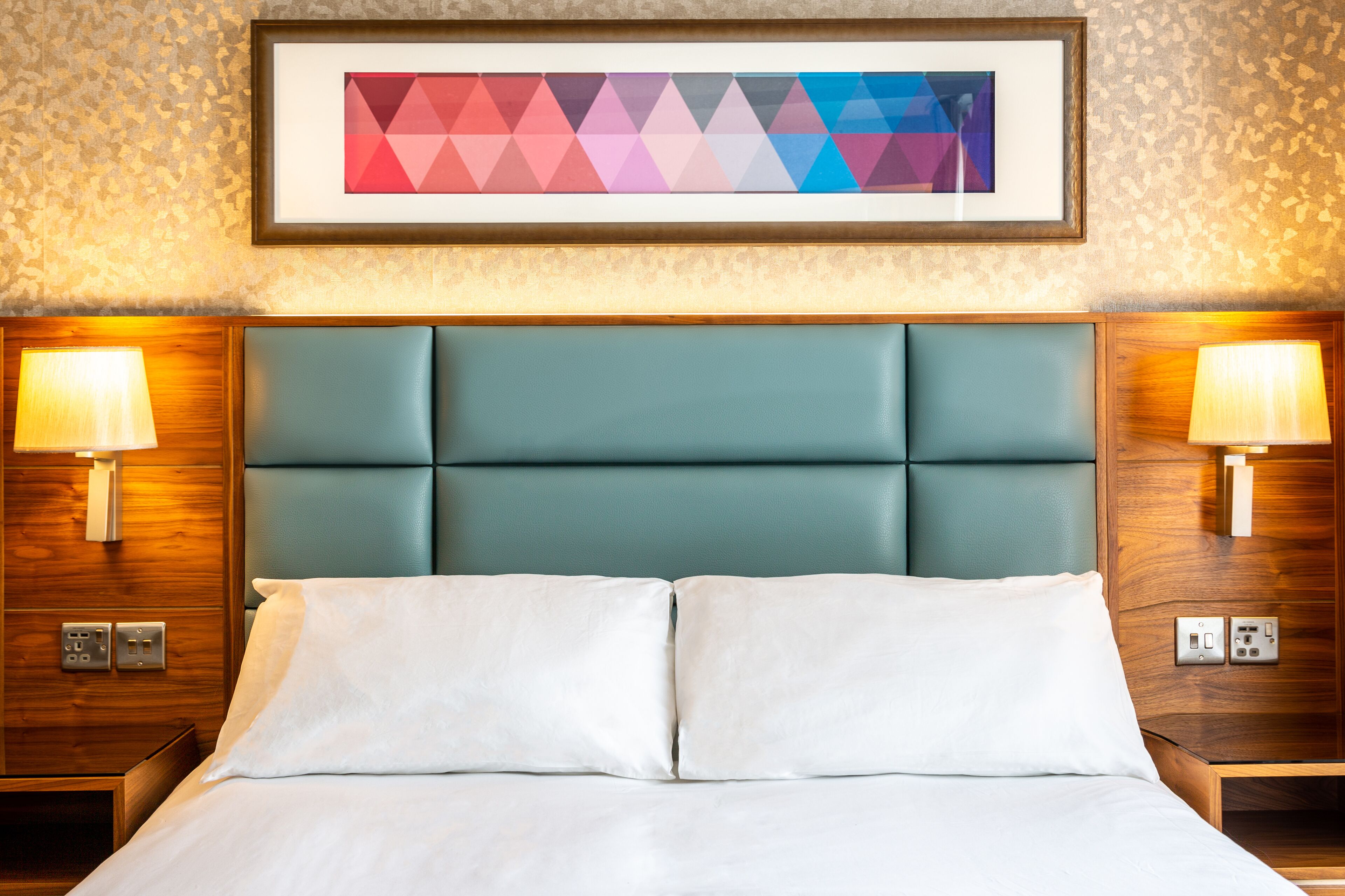A hotel room with a teal upholstered headboard, white bedding, and two bedside lamps.
