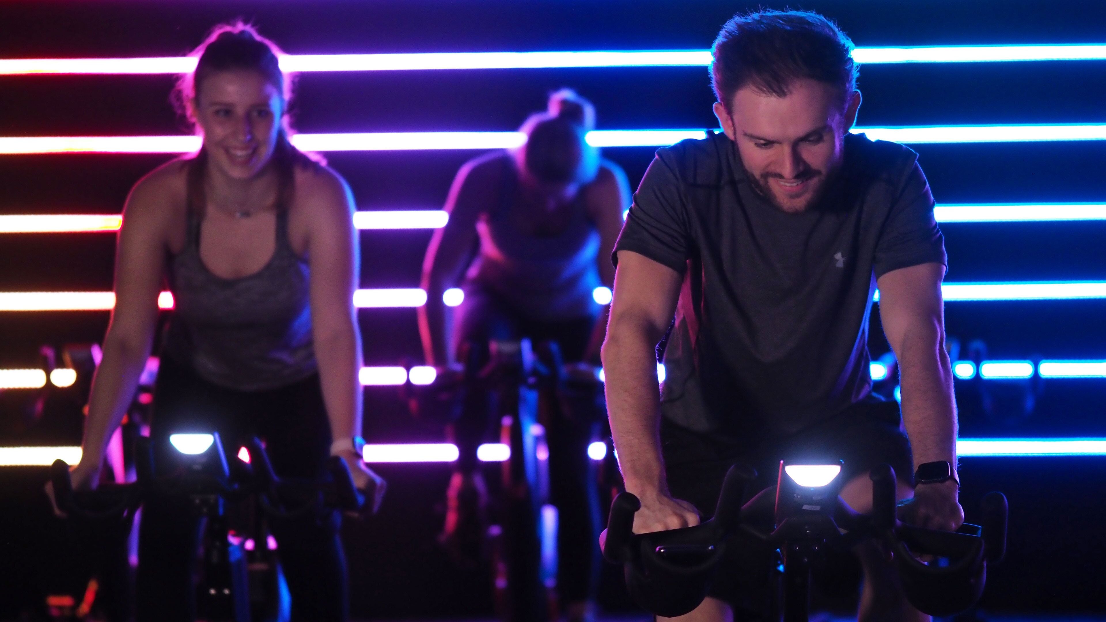 Two people on stationary bikes in a dimly lit room with horizontal neon lights.