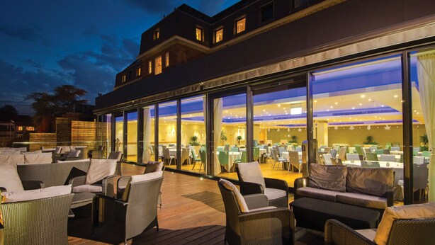 An outdoor dining area with wicker furniture and glass walls overlooking a restaurant interior. - The Lensbury Resort (Teddington)