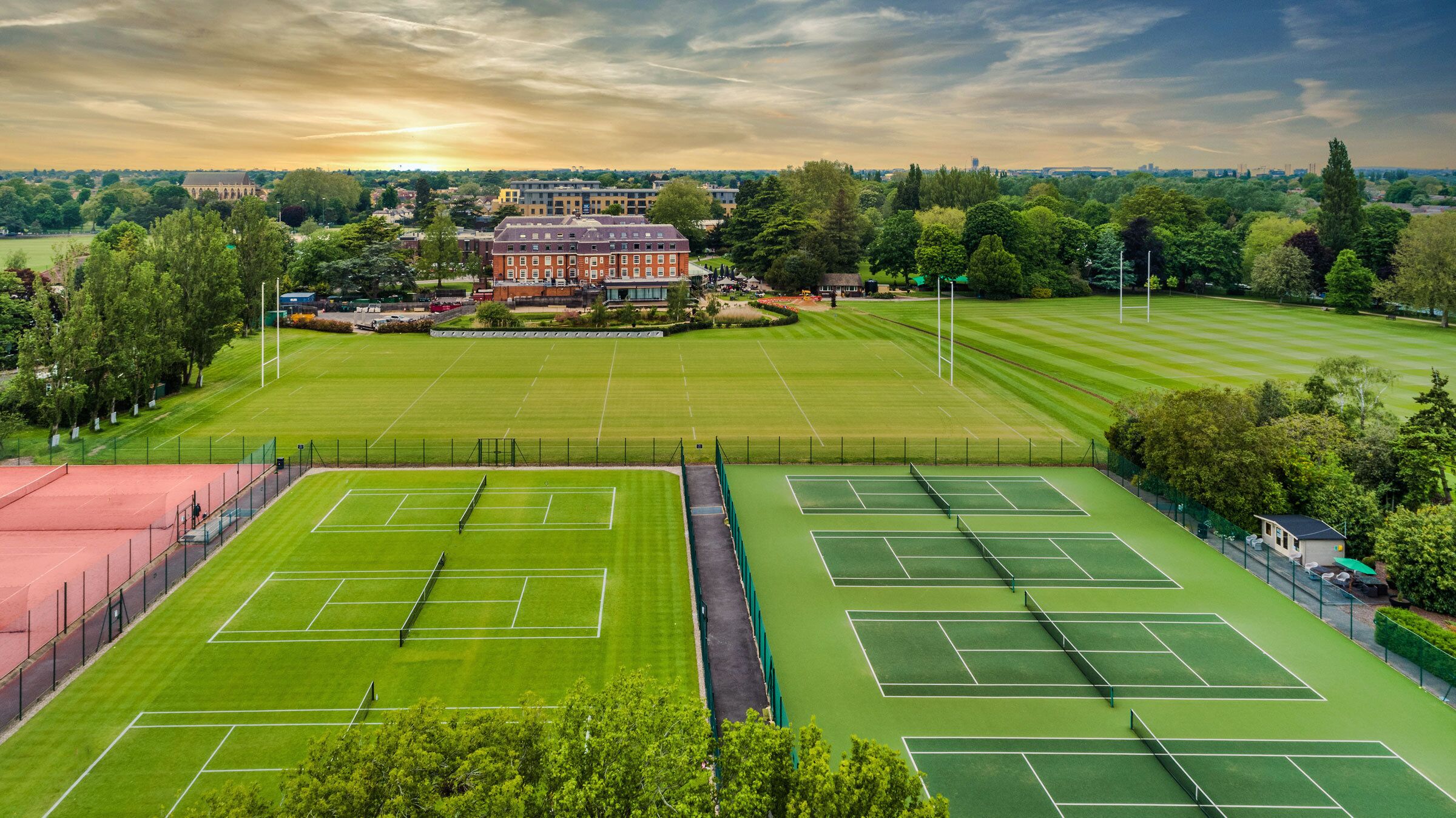 A tennis court complex with multiple courts, surrounded by trees and a large building in the background.