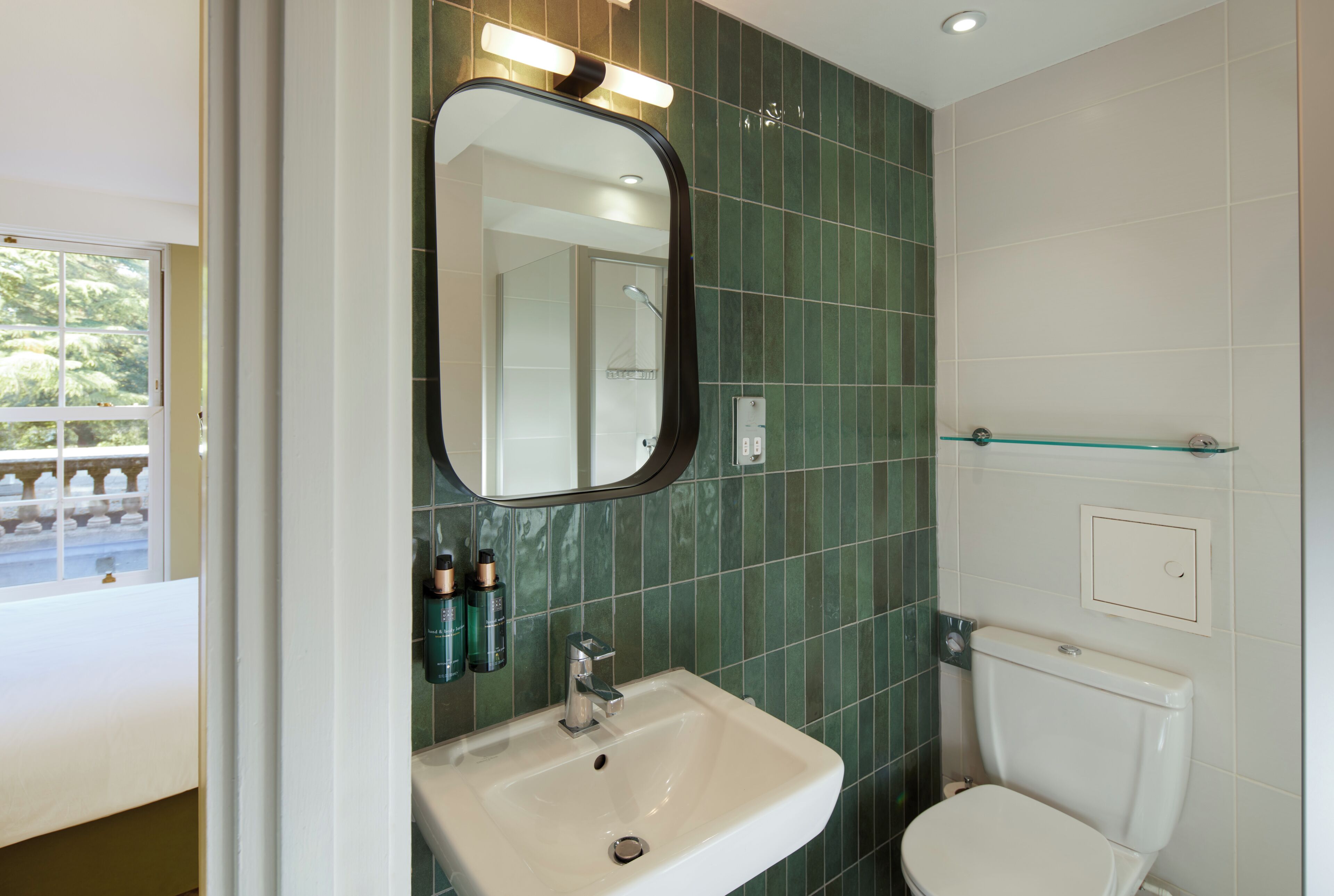 Une salle de bain avec un lavabo blanc, un miroir, des murs recouverts de carreaux verts et des toilettes blanches.
