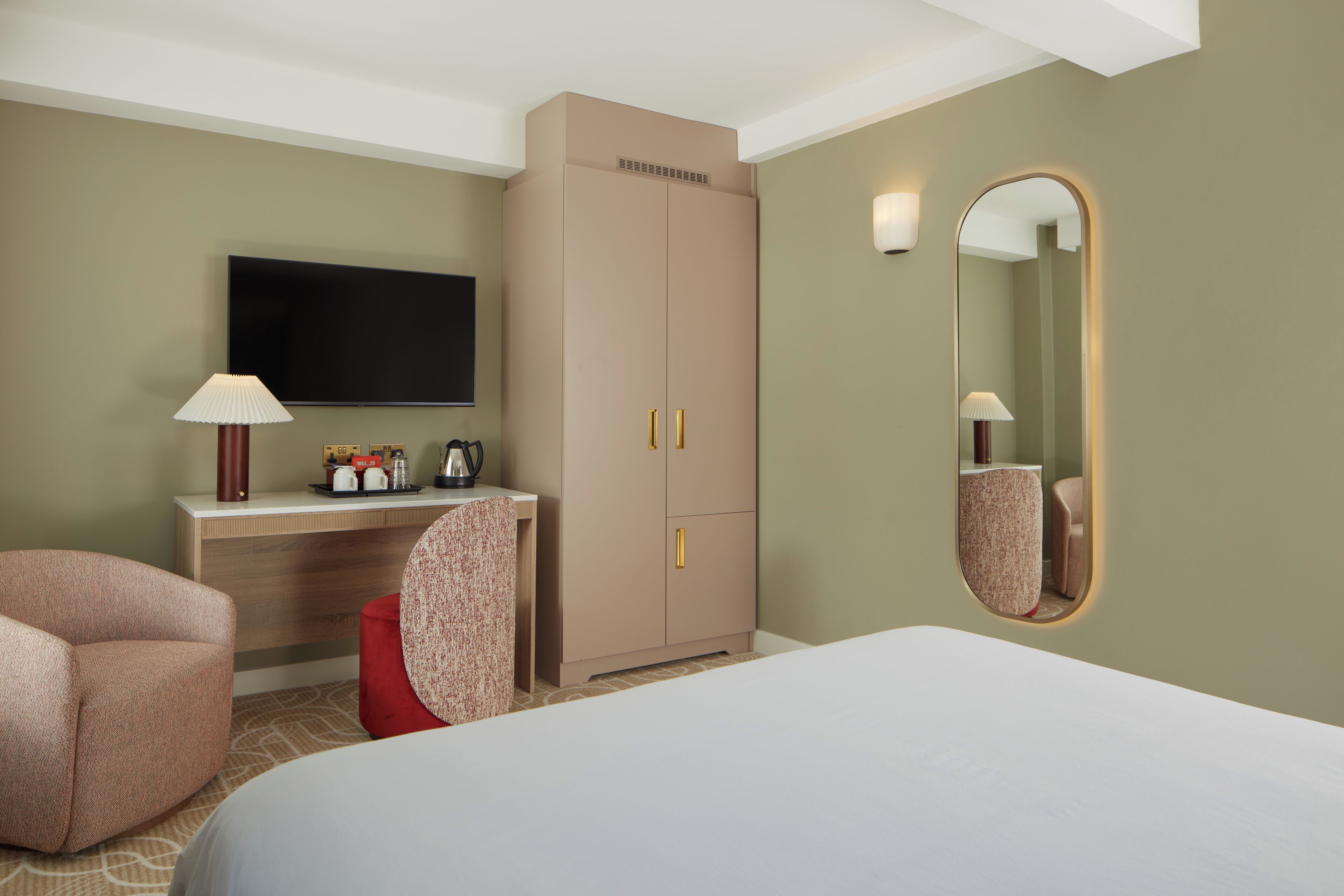 A hotel room with a bed, a desk with a flat-screen TV, a wardrobe, a chair, and a mirror.