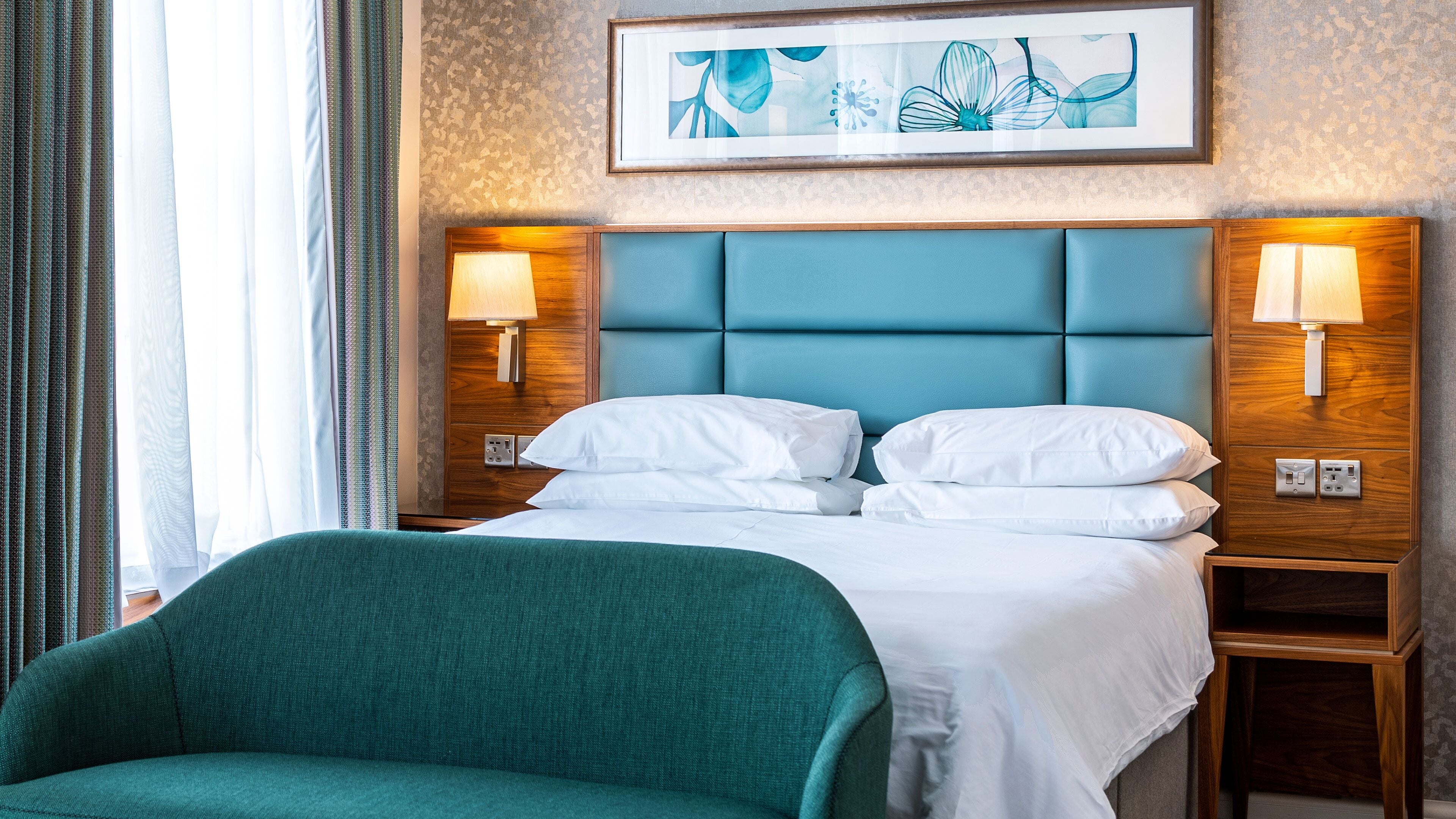 A hotel room with a large bed, two bedside tables with lamps, a green armchair, and a framed artwork on the wall.