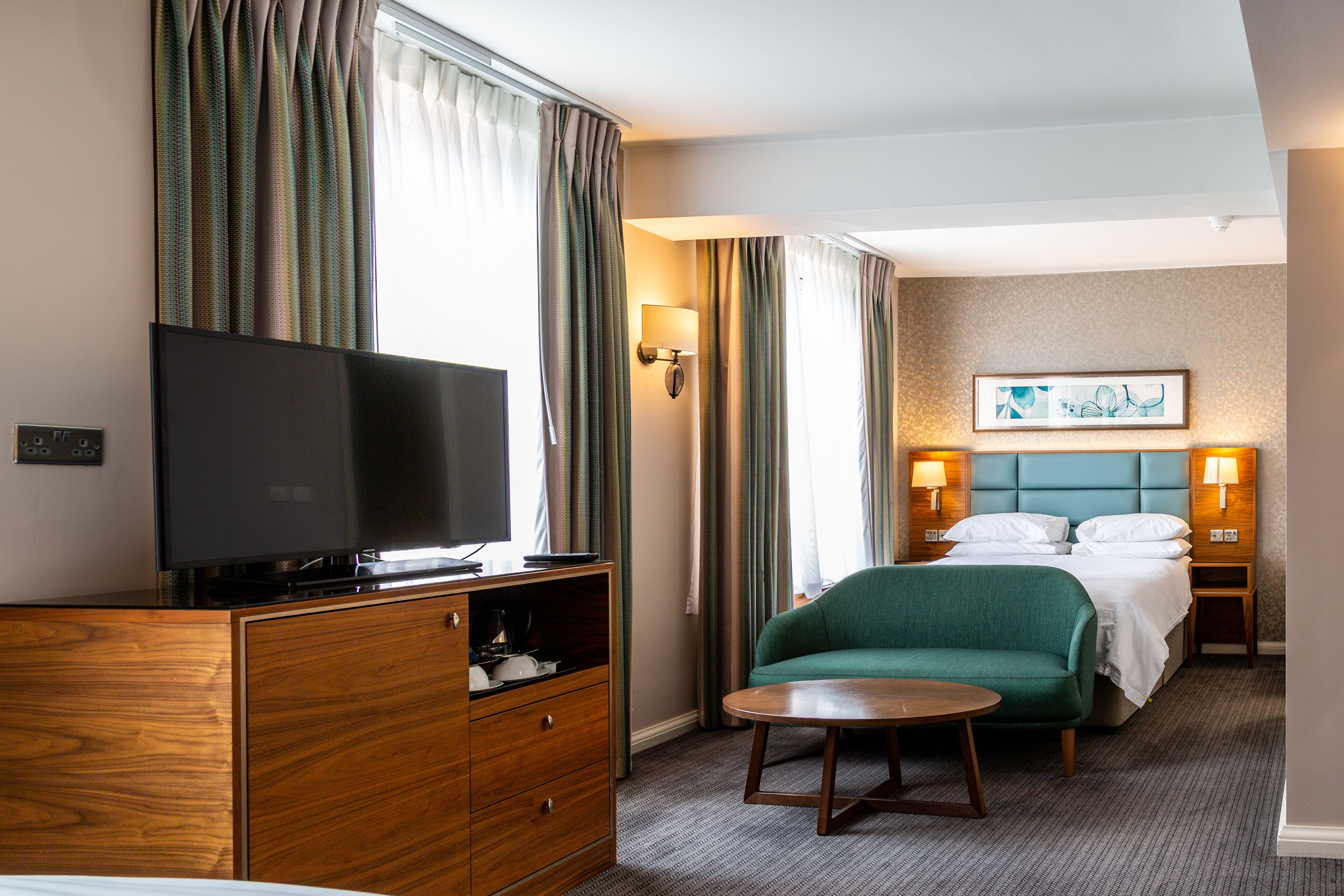 A hotel room with a television on a wooden cabinet, a green armchair, a round table, and a bed with white linens.