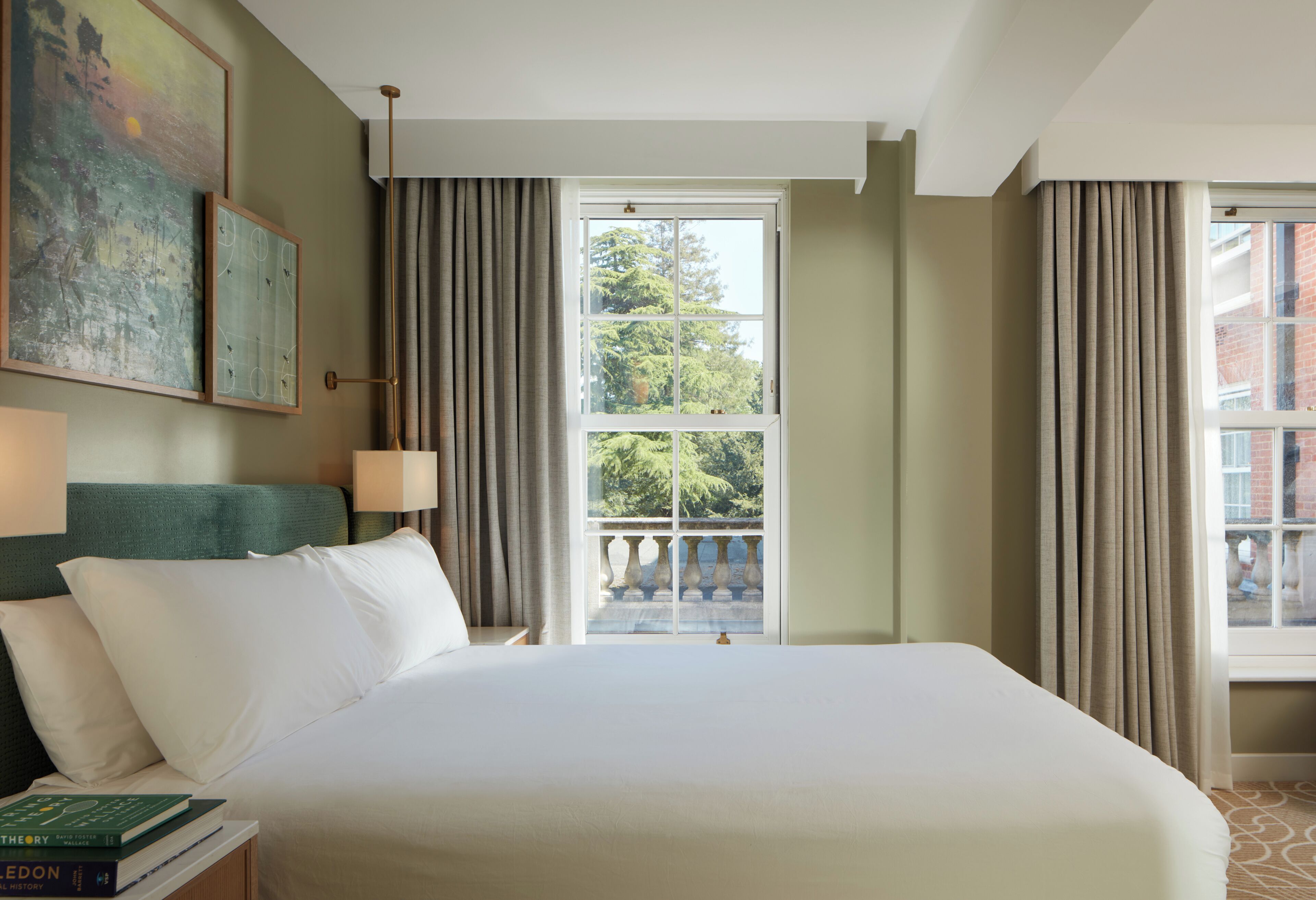 A hotel room with a large bed, a view of the outdoors, a painting on the wall, and a nightstand with books.
