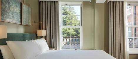 A hotel room with a large bed, a view of the outdoors, a painting on the wall, and a nightstand with books.