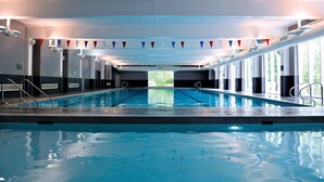 A large indoor swimming pool with clear blue water, surrounded by walls with windows and a ceiling with lights. - The Lensbury Resort (Teddington)