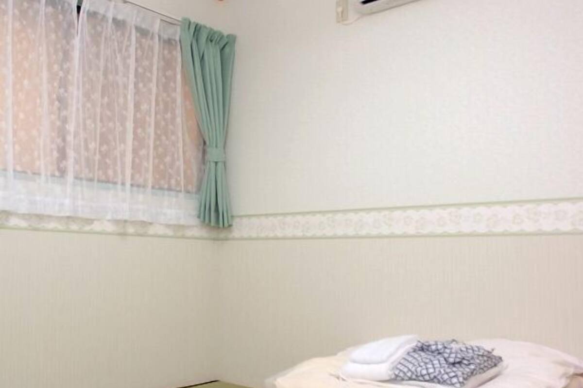 Ein Zimmer mit Tatami-Boden, einem Futonbett mit Kissen, einem Fenster mit durchsichtigen Vorhängen und einer Wand-Klimaanlage.