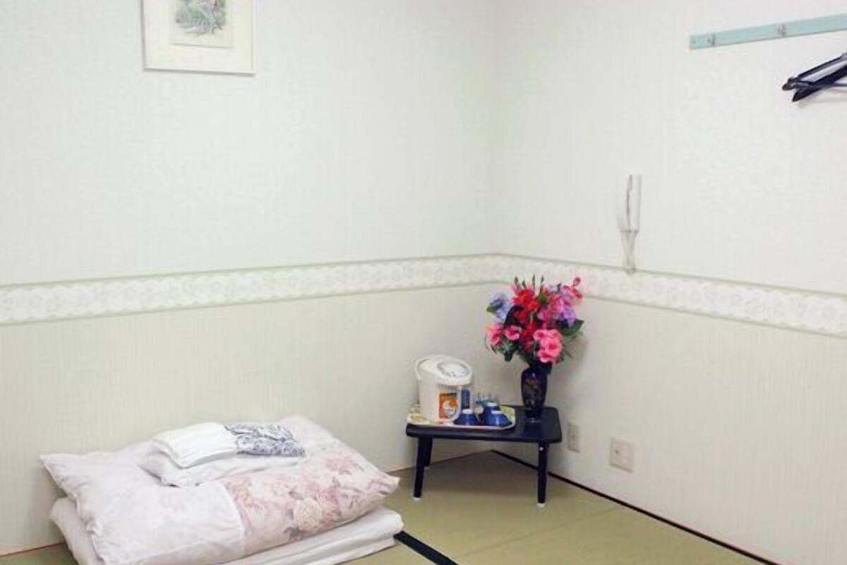Ein Zimmer mit Tatami-Bodenbelag, einem Bett mit weißer, floraler Bettdecke, einem kleinen Tisch mit Blumenvase und einem fest montierten Telefon an der Wand.