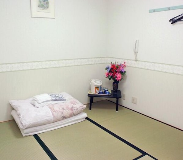 Une pièce avec un sol recouvert de tatamis, un lit avec une couette blanche à motifs floraux, une petite table sur laquelle est posé un vase de fleurs et un téléphone fixé au mur.