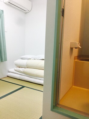 Ein Zimmer mit Tatami-Boden, einem Bett mit weißer Bettwäsche, einer Klimaanlage an der Wand und einem Holztürrahmen.