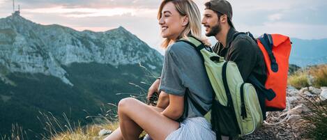 Two people sitting on a rocky outcrop with backpacks, overlooking a mountainous landscape.