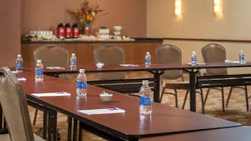 Un salón de conferencias con mesas largas, sillas, botellas de agua y una pared decorativa con un jarrón y velas.