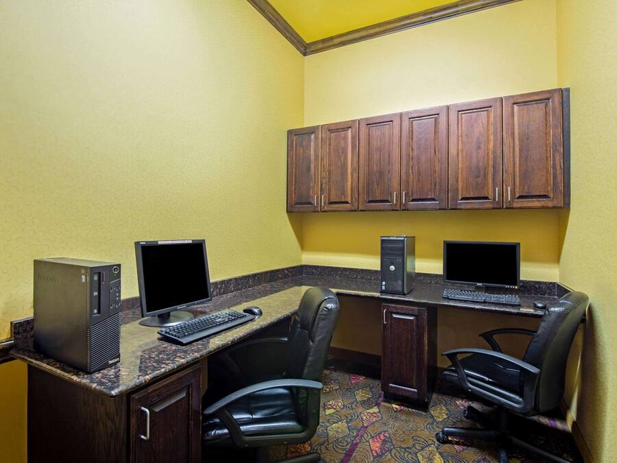 A hotel room with two desks, each with a computer, keyboard, and mouse. There are two chairs, one at each desk. The room has wooden cabinets above the desks and a patterned carpet on the floor.