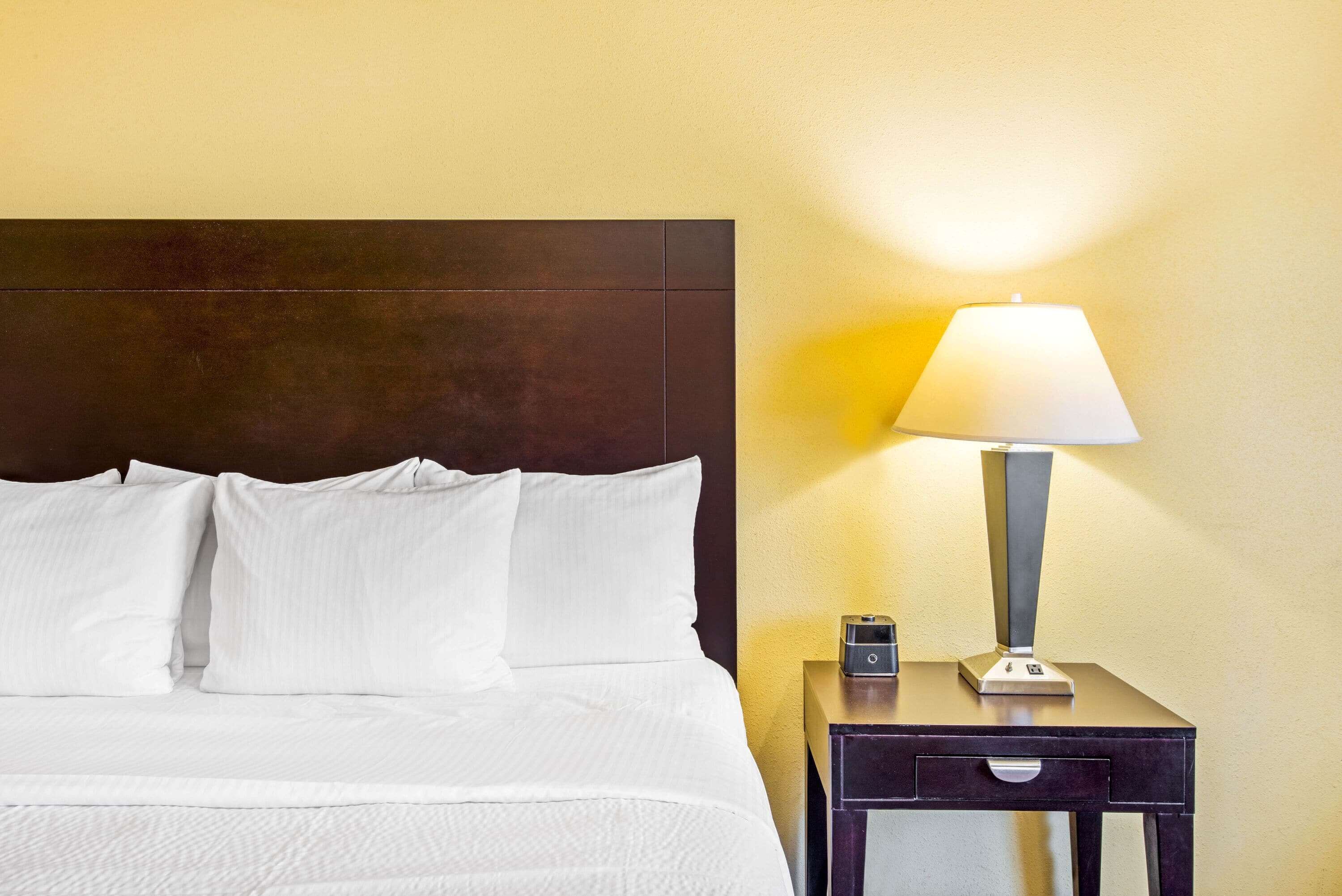 A neatly made bed with white linens and pillows, a bedside table with a lamp, and a small electronic device on the table.