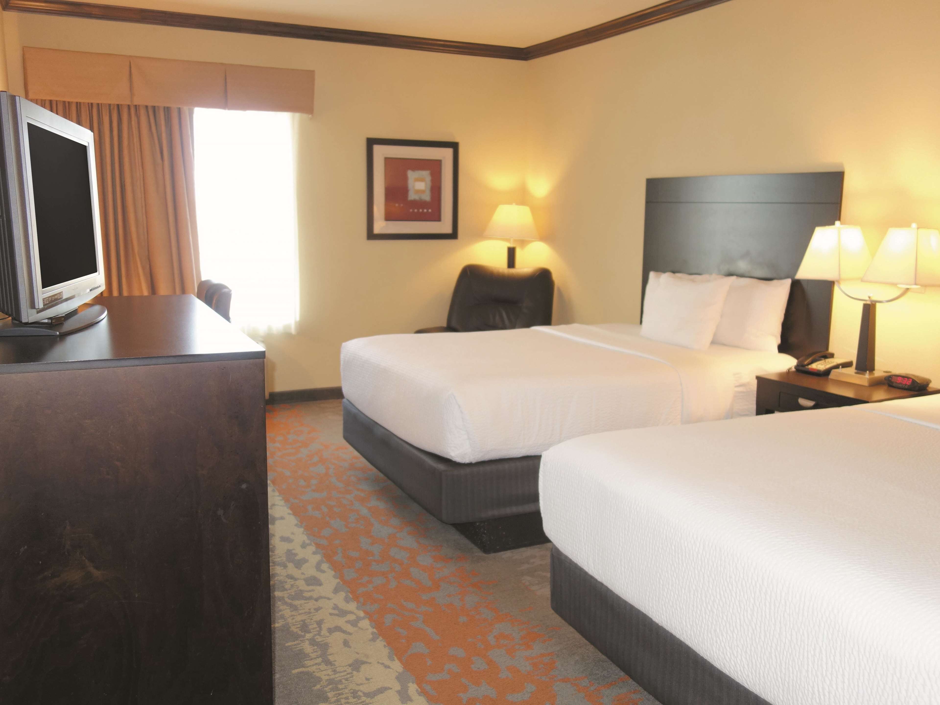 A hotel room with two beds, a desk with a television, a framed picture on the wall, and a lamp on the nightstand.
