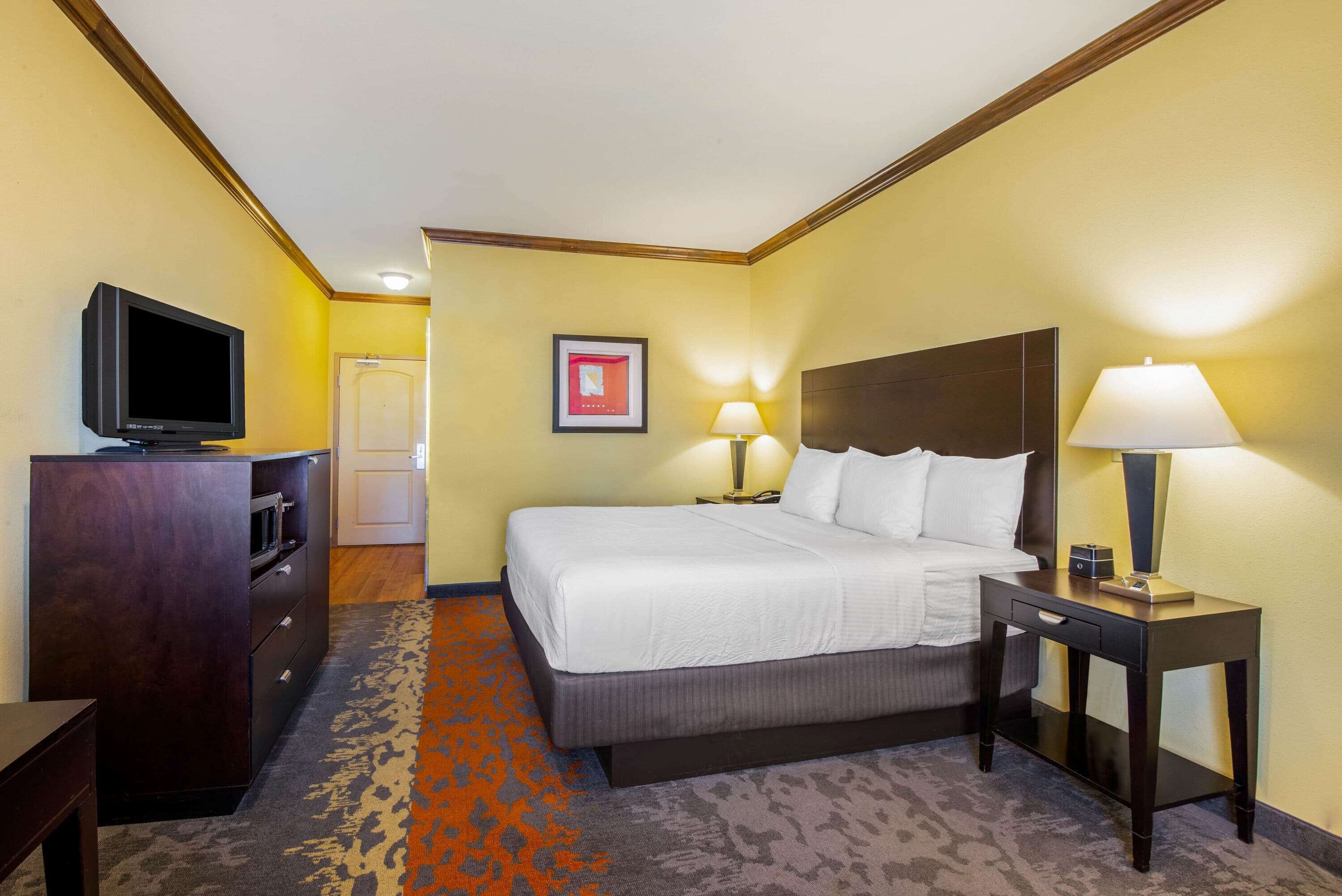A hotel room with a bed, a desk with a lamp, a television, and a door.