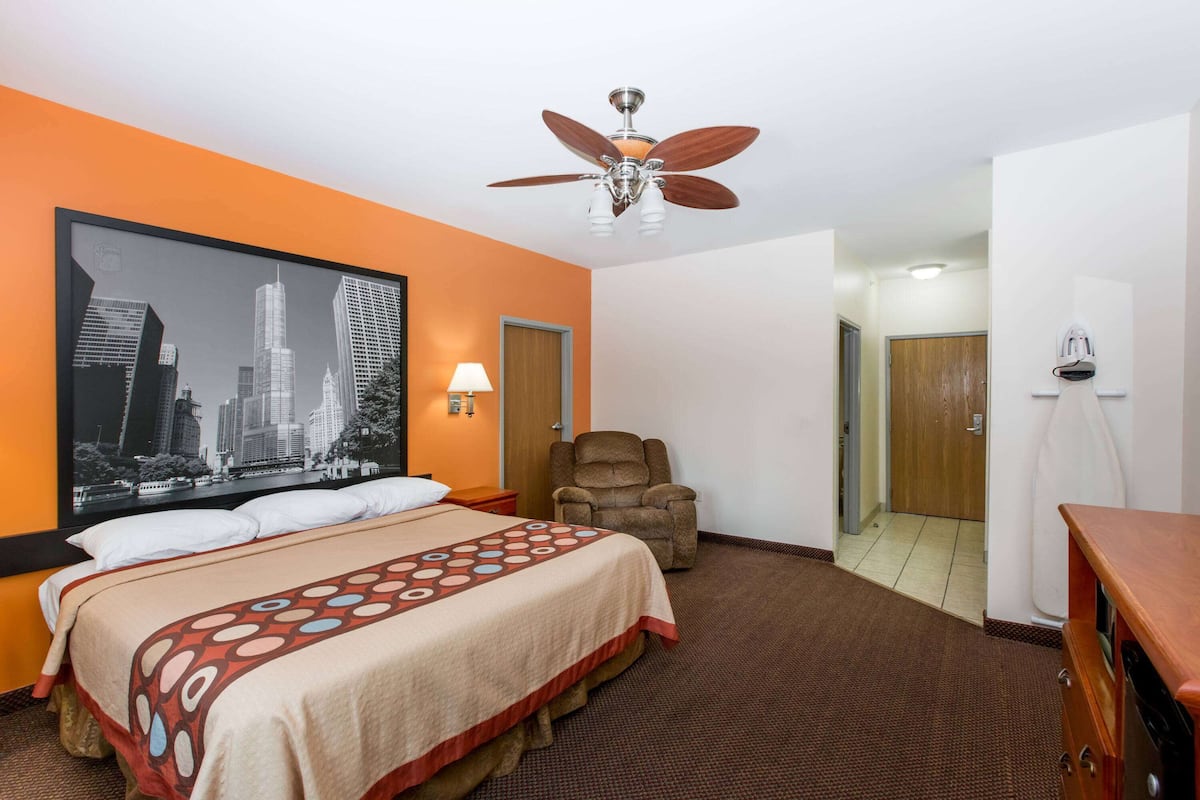 Habitación de hotel con cama, una silla, ventilador de techo y un cartel con un paisaje urbano en la pared.