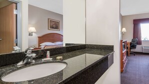 Een hotelbadkamer met een grote spiegel, een wastafel en een bed zichtbaar op de achtergrond.
