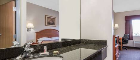 A hotel bathroom with a large mirror, a sink, and a bed visible in the background.