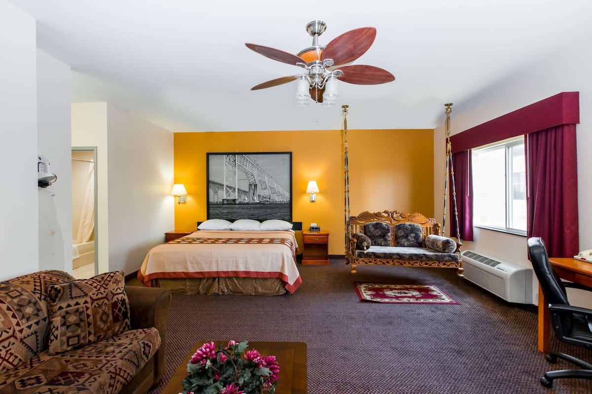 Habitación de hotel con cama, sofá, escritorio, silla, ventilador de techo, calentador de pared y ventana con cortinas.