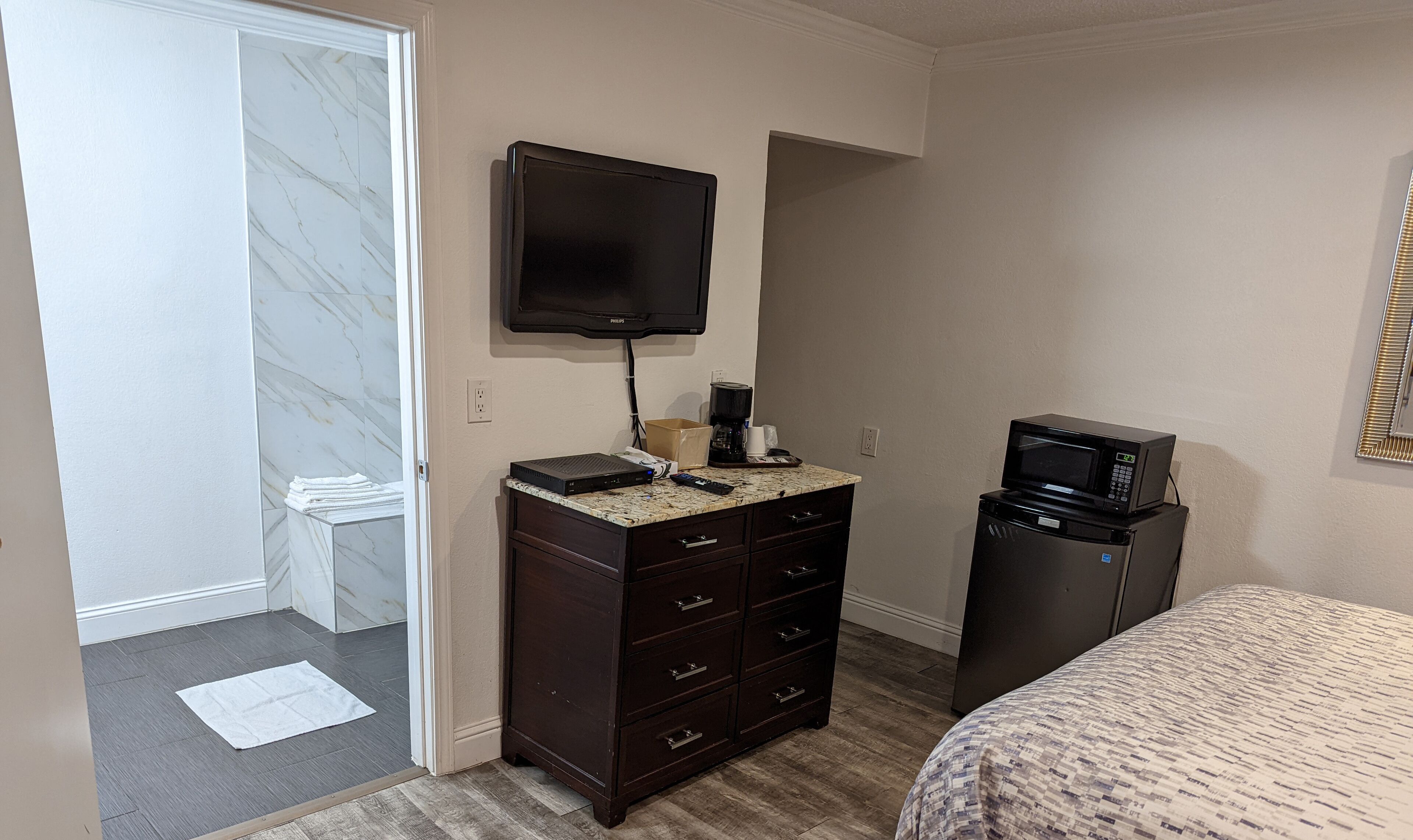 Una habitación de hotel con una cama, un aparador con cajones, un televisor montado en la pared, un microondas y una cafetera.