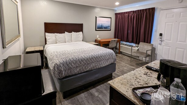 A hotel room with a large bed, a desk, a chair, a coffee maker, and a TV. - Deluxe Inn (South San Francisco)